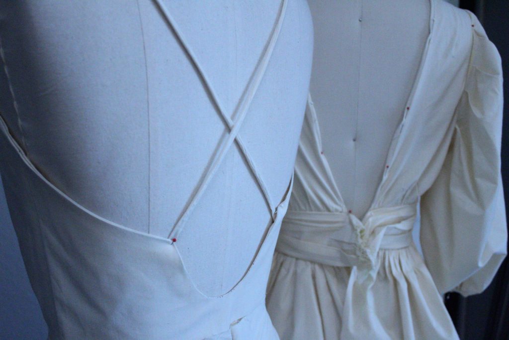 Muslin test garments on dress forms one low back with thin crossing straps. One open back with balloon sleeves